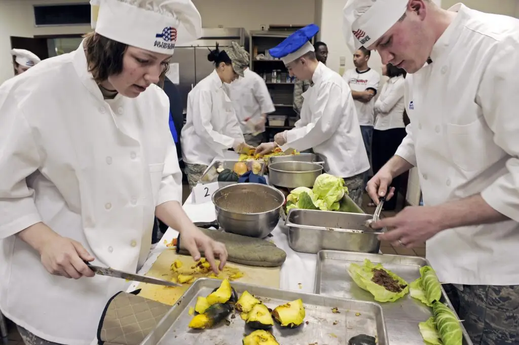 "Several chefs in white uniforms and various styles of hats are busily preparing food in a commercial kitchen. They are focused on chopping and peeling vegetables like squash and cabbage on stainless steel countertops. In the background, other cooks are actively engaged in cooking, with pots on stoves and ingredients laid out around them. The scene is bustling with culinary activity, indicative of a professional kitchen environment."